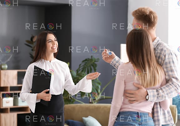 A woman in a white shirt is holding a clipboard and talking to two other people in a room with a couch and a plant.