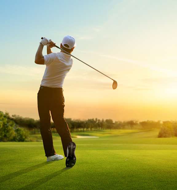 A golfer is captured in mid-swing on a golf course at sunset.