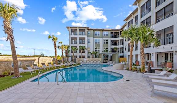 A large swimming pool surrounded by palm trees and lounge chairs in front of a multi-story apartment building.