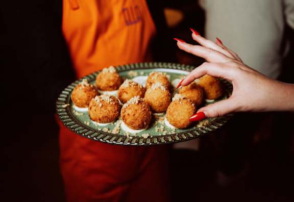 A person in an orange shirt is holding a plate of round, golden-brown food items.