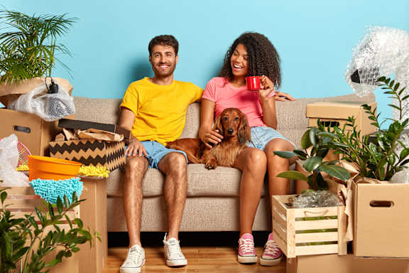 A man and woman sitting on a couch with a dog between them, surrounded by moving boxes and plants.