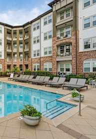 A large swimming pool surrounded by lounge chairs and potted plants in front of a multi-story apartment building.