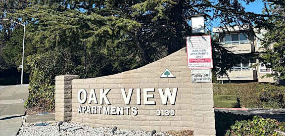 A sign for Oak View Apartments stands in front of a large tree.