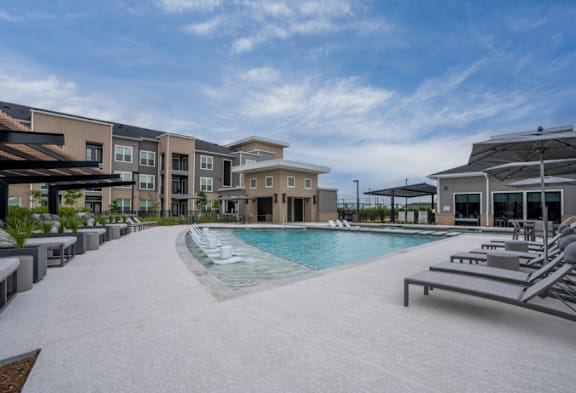 A swimming pool surrounded by sun loungers and umbrellas in front of apartment buildings.