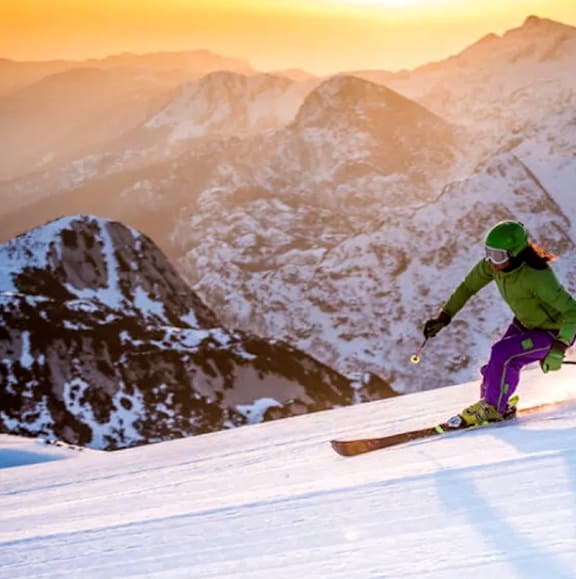 A skier in a green jacket and purple pants is skiing down a snowy mountain.