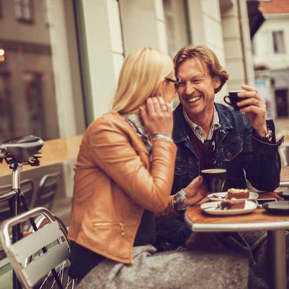 A man and a woman are sitting at a table outside a cafe, with the woman talking on the phone and the man holding a cup.