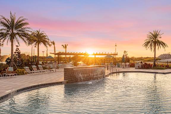 A sunset view of a pool with a waterfall and palm trees.
