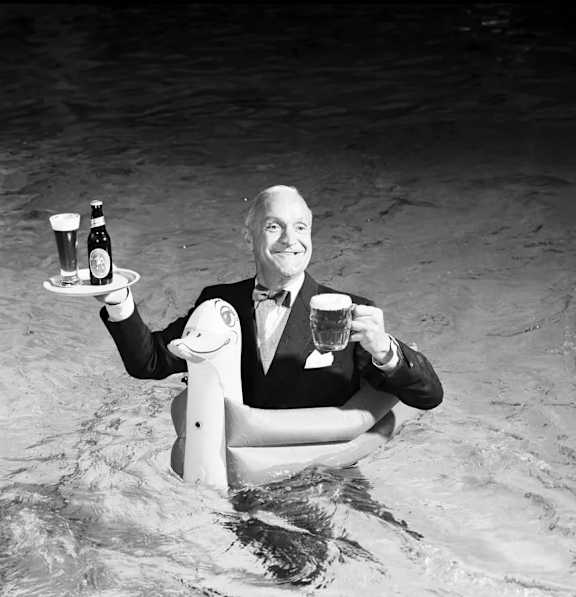 A man in a tuxedo is sitting in the water holding a tray with two beers and a stuffed animal.