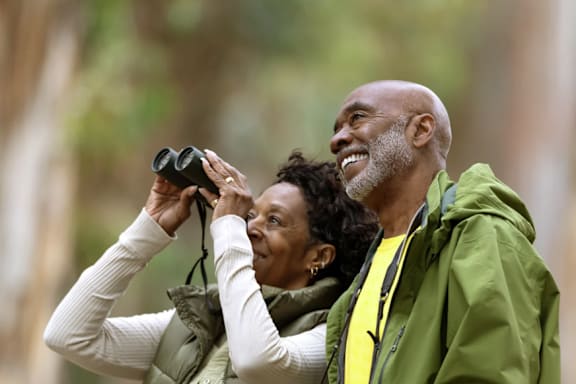 A man and a woman are looking through binoculars in a wooded area.