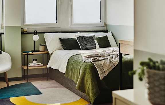 A bedroom with a bed, a chair, a table with a lamp, and a potted plant.