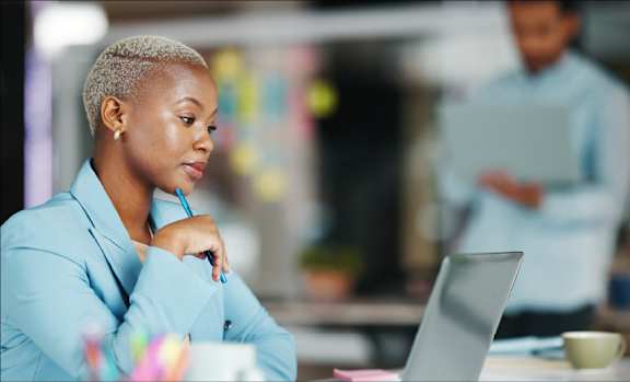 A woman in a blue blazer is working on a laptop in an office environment.