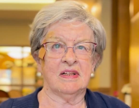 An elderly woman with glasses and a blue sweater is speaking.