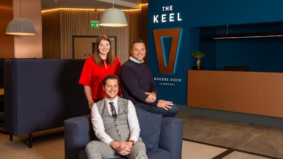two men and a woman sitting on a blue couch in front of the keel at The Keel Apartments, Liverpool, L3 4GE