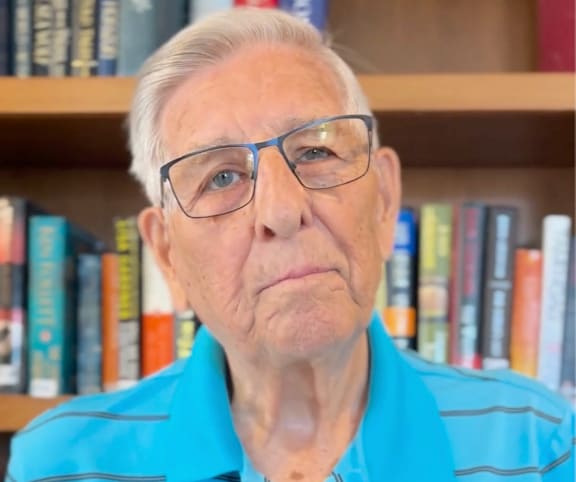 An older man with glasses and a blue striped shirt is standing in front of a bookshelf.