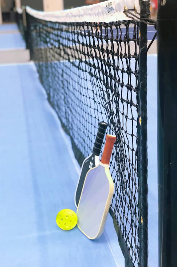 A tennis racket and ball are leaning against a tennis net.