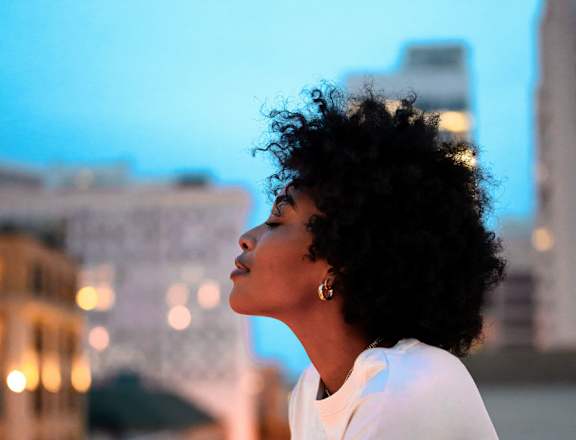 A woman with curly hair is standing on a balcony looking at the city lights.