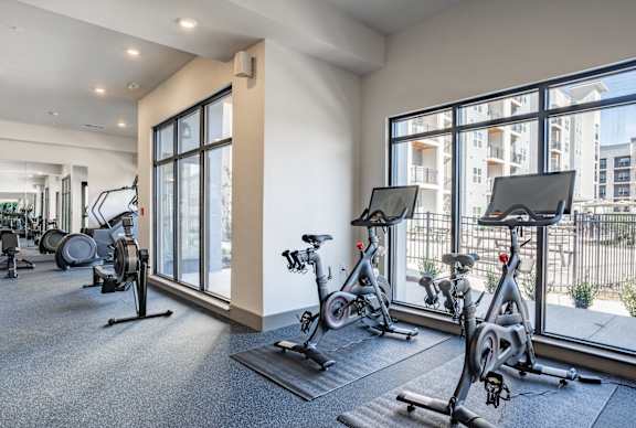 A gym with exercise bikes and a view of a building outside the window.