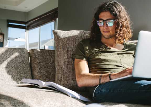 A man with long hair is sitting on a couch, reading a book and using a laptop.