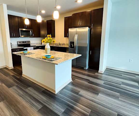 A large, modern kitchen with stainless steel appliances, kitchen utensils, marble countertops with lots of room, a kitchen island with two places set, a vase with sunflowers, and large dark wooden cabinets at Royster Commons Apartment Homes. 