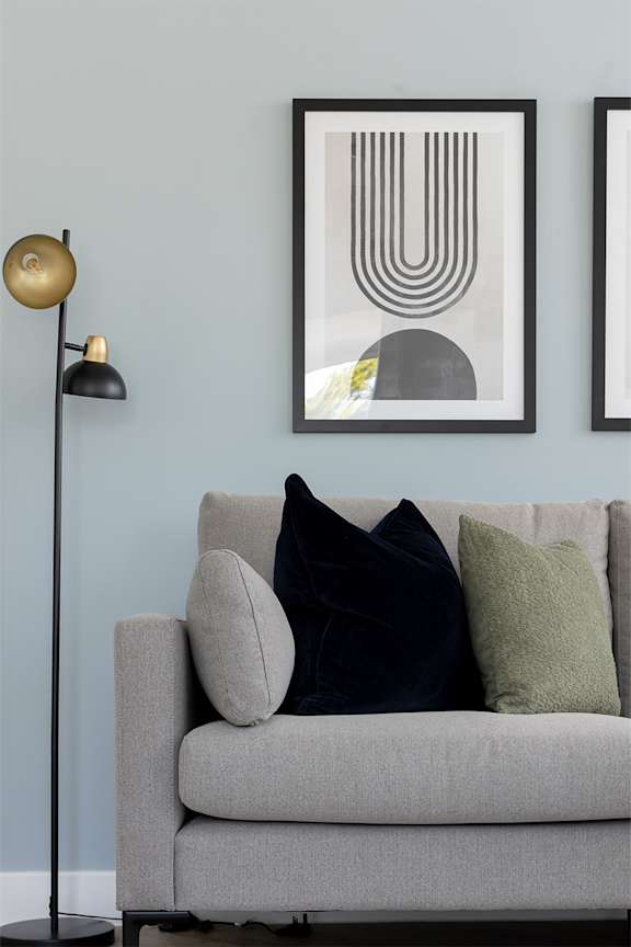 A grey couch with two dark and one light green pillow is in front of two framed artworks on a wall.