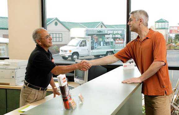 Two men shake hands in front of a window with a garage sign in the background.