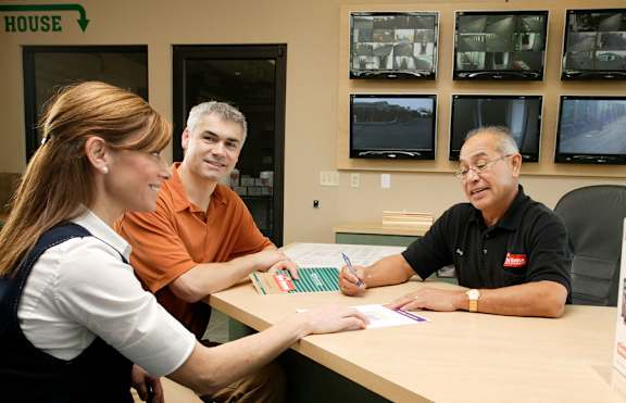 A woman in a white shirt and vest is sitting at a desk with two men, one in an orange shirt and the other in a black shirt, in front of a wall with multiple monitors.