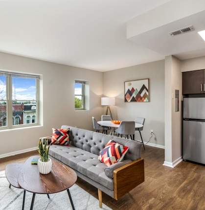 A living room with a grey couch, a table, a chair, a potted plant, and a window.