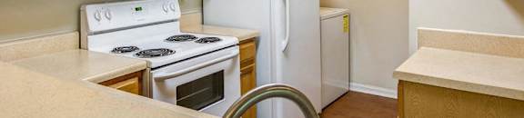 L Shaped Kitchen with Blonde Cabinets Tan Counters and White Appliances and Utility Closet with Washer Visible in the Background