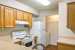 L Shaped Kitchen with Blonde Cabinets Tan Counters and White Appliances and Utility Closet with Washer Visible in the Background