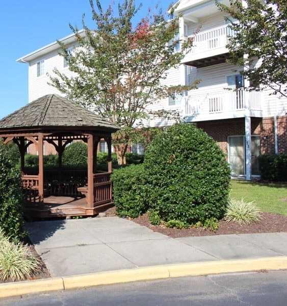 A gazebo is surrounded by bushes and trees in front of a white building at Greenbrier Seniors Apartments, Chesapeake, VA 23320  .