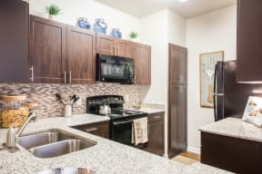 North Austin, TX Apartments - Kitchen With Granite-Style Countertops, Wood-Style Cabinets, Modern Appliances, Microwave, Oven, Dual Sink, Fridge, Tile Back-Splash, And Wood-Style Flooring.