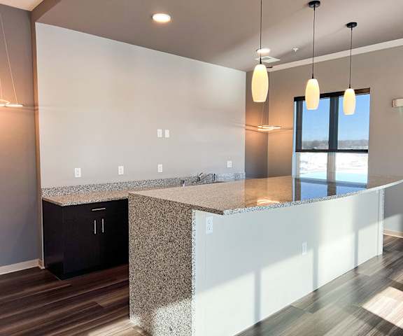 An empty, modern kitchen with granite countertops, unique lighting, and a window at Royster Commons Apartment Homes. 