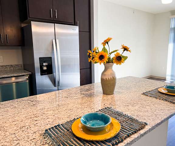 A granite kitchen counter with a vase full of sunflowers in it and two places set with a stainless-steel refrigerator in the background at Royster Commons Apartment Homes. 