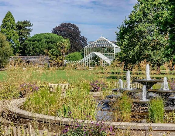 A garden with a fountain in the foreground and a greenhouse in the background at Origins Apartments, Cambridge, CB3 0UX