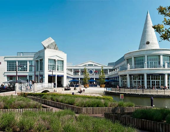 A modern building with a white pyramid-shaped structure on top is situated next to a body of water with people walking around at UnionView Apartments, Dartford DA2 6GZ