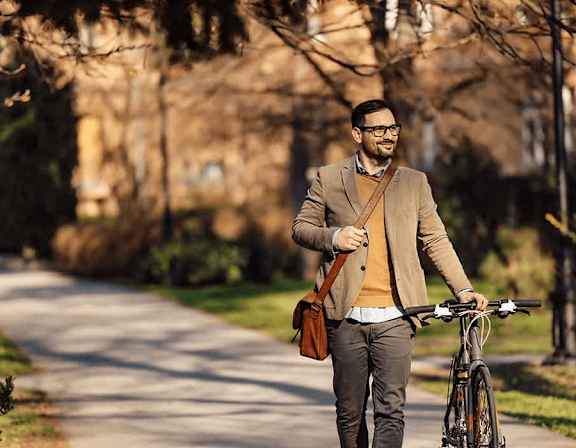 A man in a brown jacket and glasses is walking with a bicycle and holding a brown bag. at Origins Apartments, Cambridge