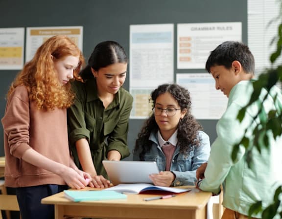 Four students are gathered around a table, looking at a laptop screen and discussing the information displayed. at The Woodlands Apartments, Milton Keynes, MK5 6GS