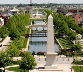 A monument stands in a park with a river and a bridge in the background.