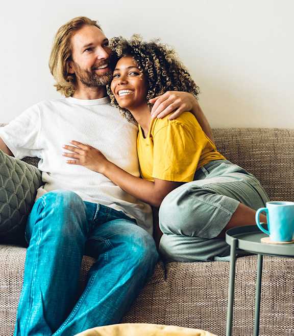 A couple is sitting on a couch, the man is wearing a white t-shirt and blue jeans, and the woman is wearing a yellow top and grey pants.