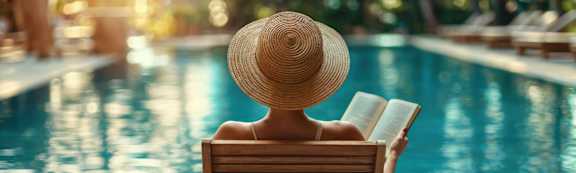 A person in a straw hat is sitting on a lounge chair by a pool.