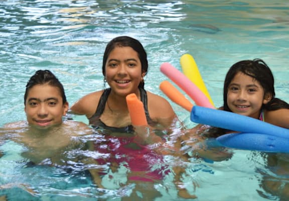 Three girls are in a pool with pool noodles.