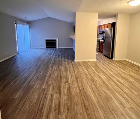 an empty living room with a fireplace and a kitchen in the background