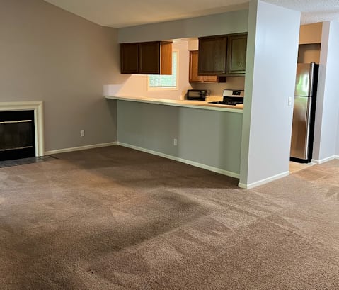 an empty living room with a fireplace and a kitchen in the background