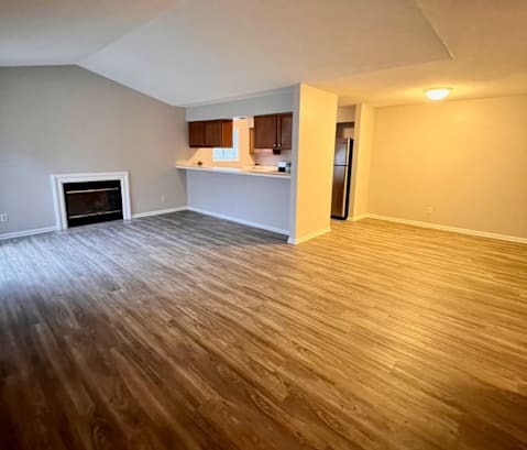 an empty living room with a fireplace and a kitchen in the background