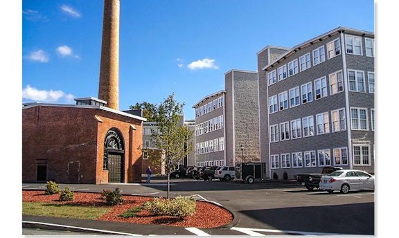 A brick building with a tall chimney sits next to a modern building with a parking lot in front.