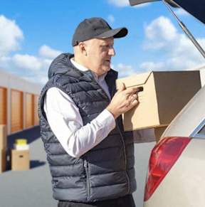 A man in a black vest and cap is loading a cardboard box into the trunk of a beige car.