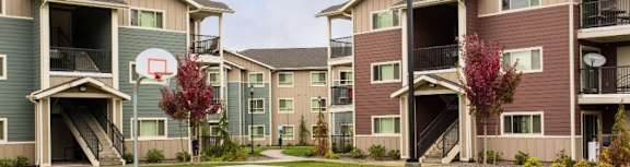 A basketball court is in the middle of a grassy area in front of apartment buildings.