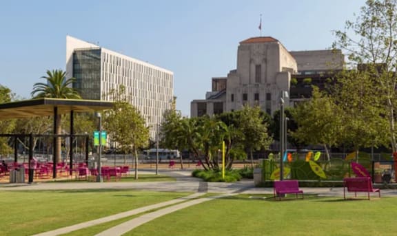 A park with a grassy area, benches, and trees in front of a building with a flag on top.