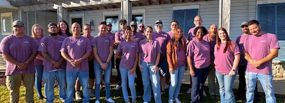 A group of people wearing purple shirts are standing in front of a house.