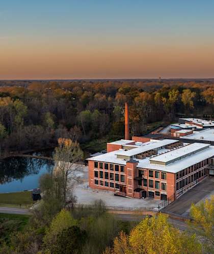 A large building with a tall chimney is surrounded by trees and a body of water.